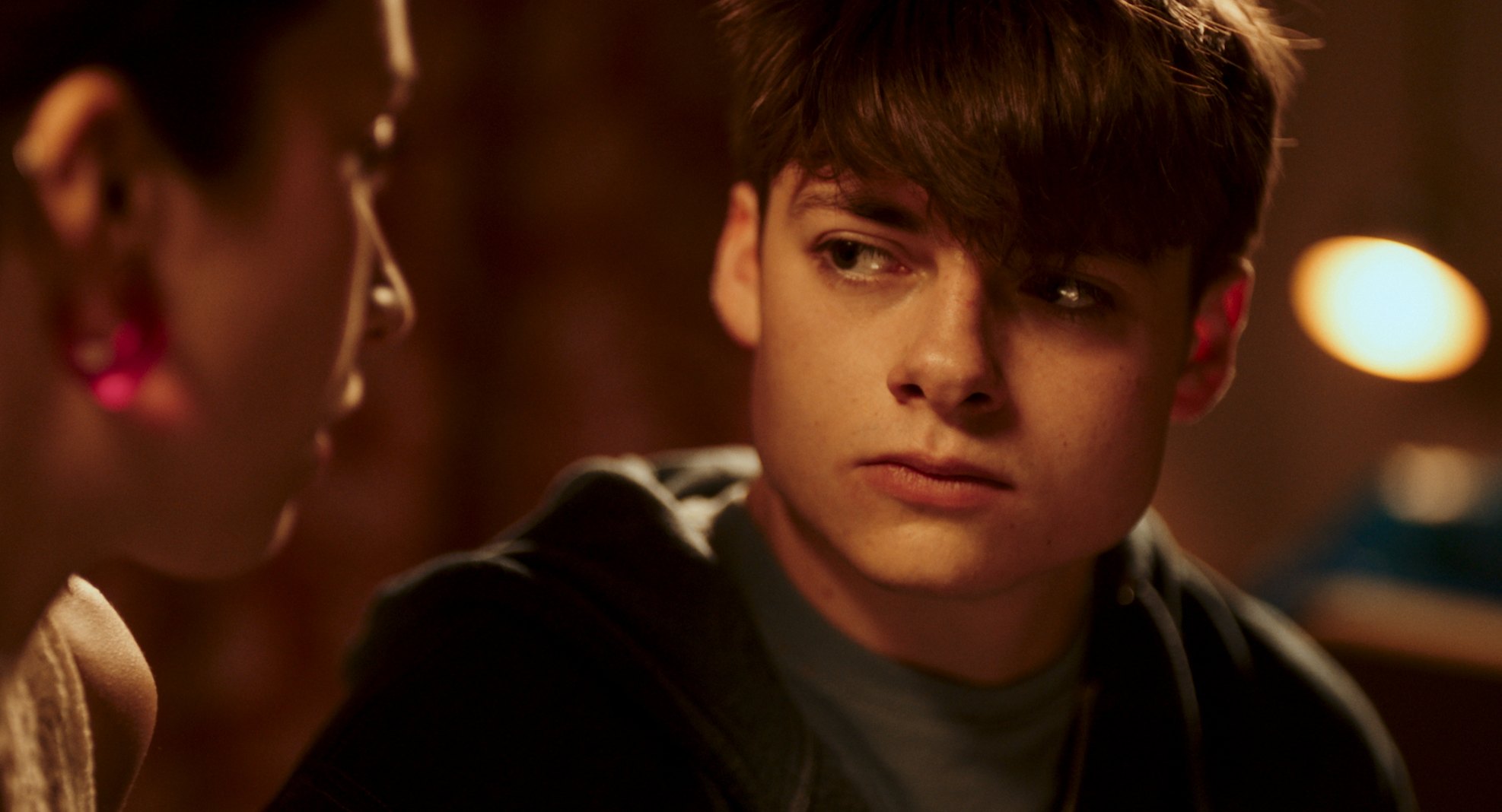 A young man with a floppy fringe glances sideways in warm, low light.