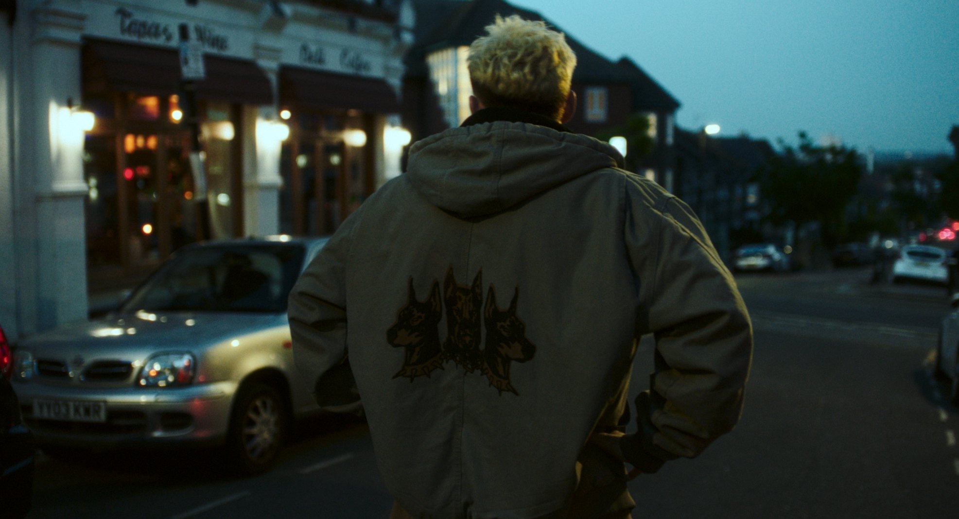 A man with bleached hair seen from behind, walking down a dusk street in a jacket with a three-dog patch.