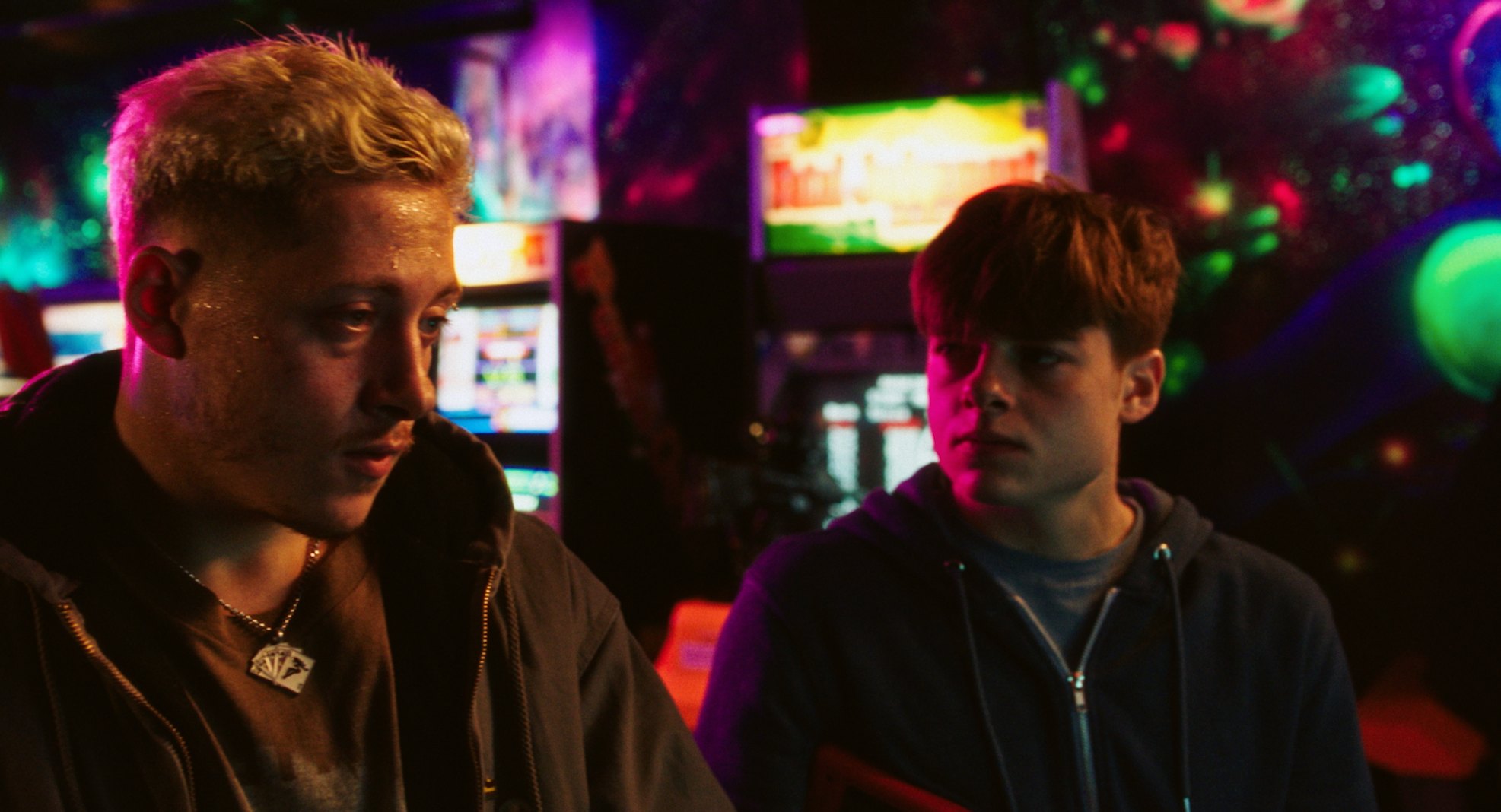 Two young men in a neon-lit arcade, one looking down, the other watching him.