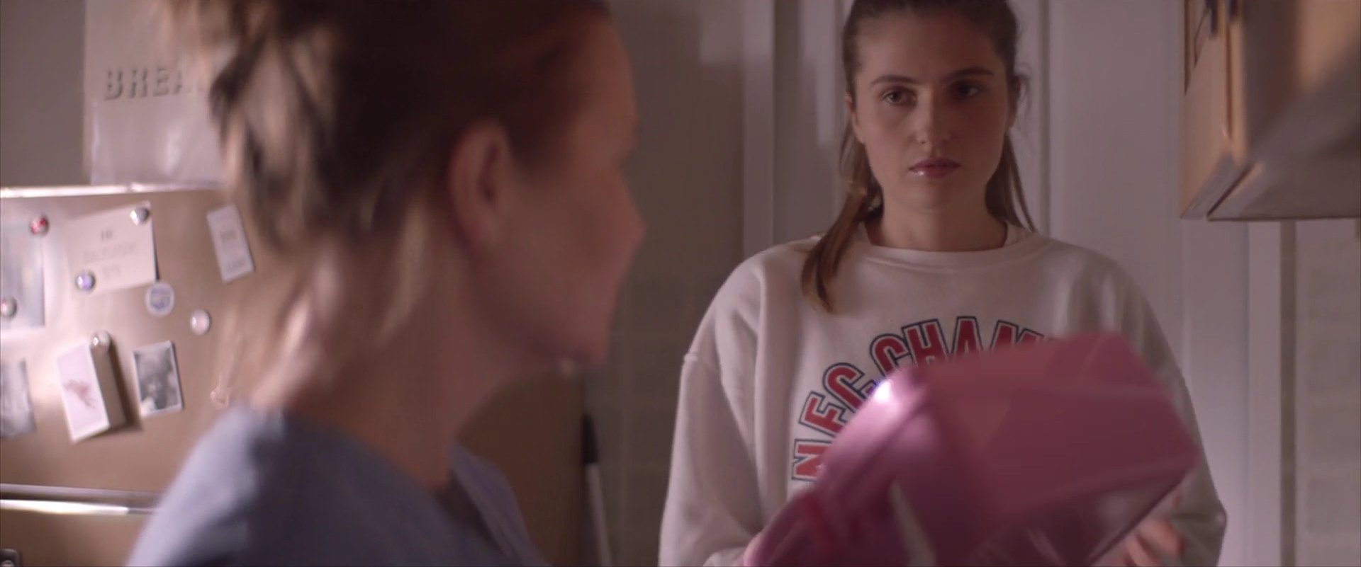 A young woman in a white sweatshirt stands in a kitchen holding a pink container, watching a woman in the foreground.