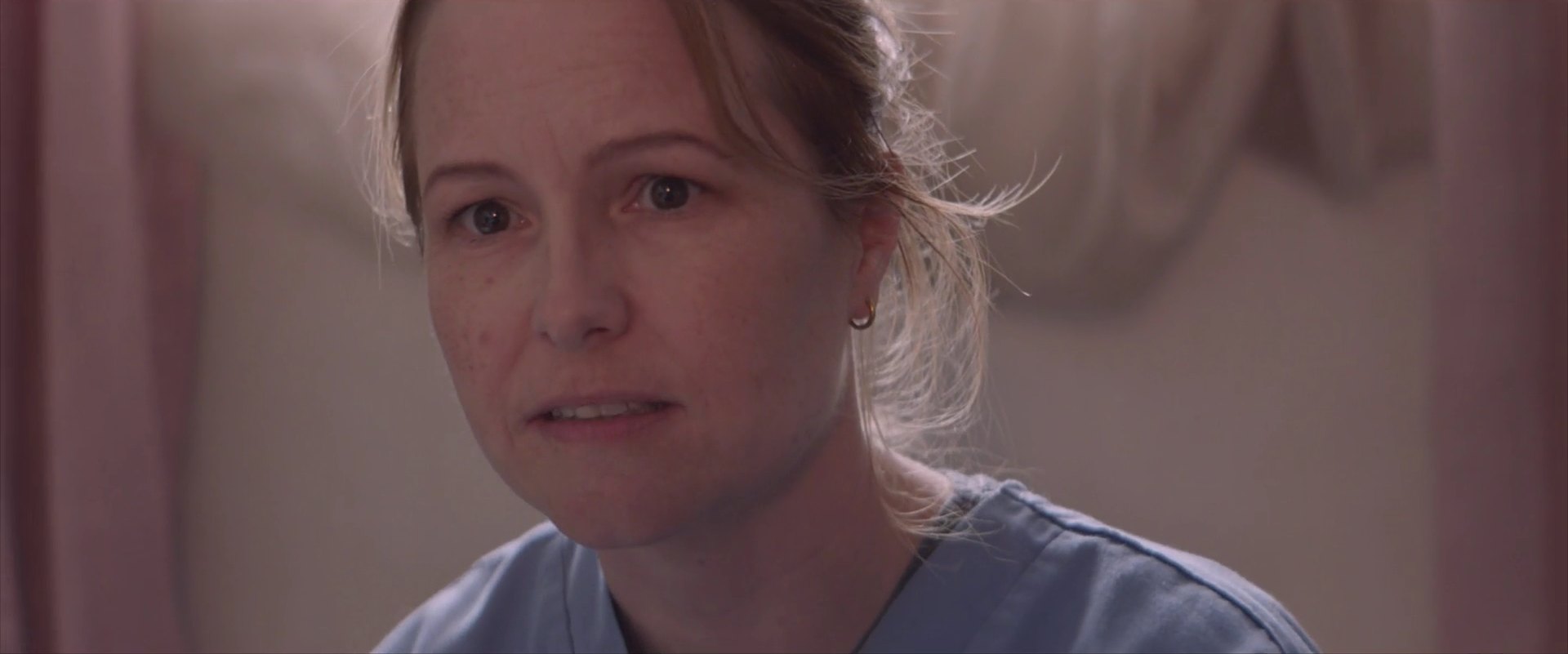 Close-up of a woman in blue scrubs with her hair tied back, speaking.