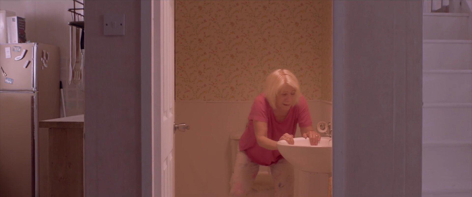 A woman in pink leans over a bathroom sink, seen through a doorway from the kitchen.