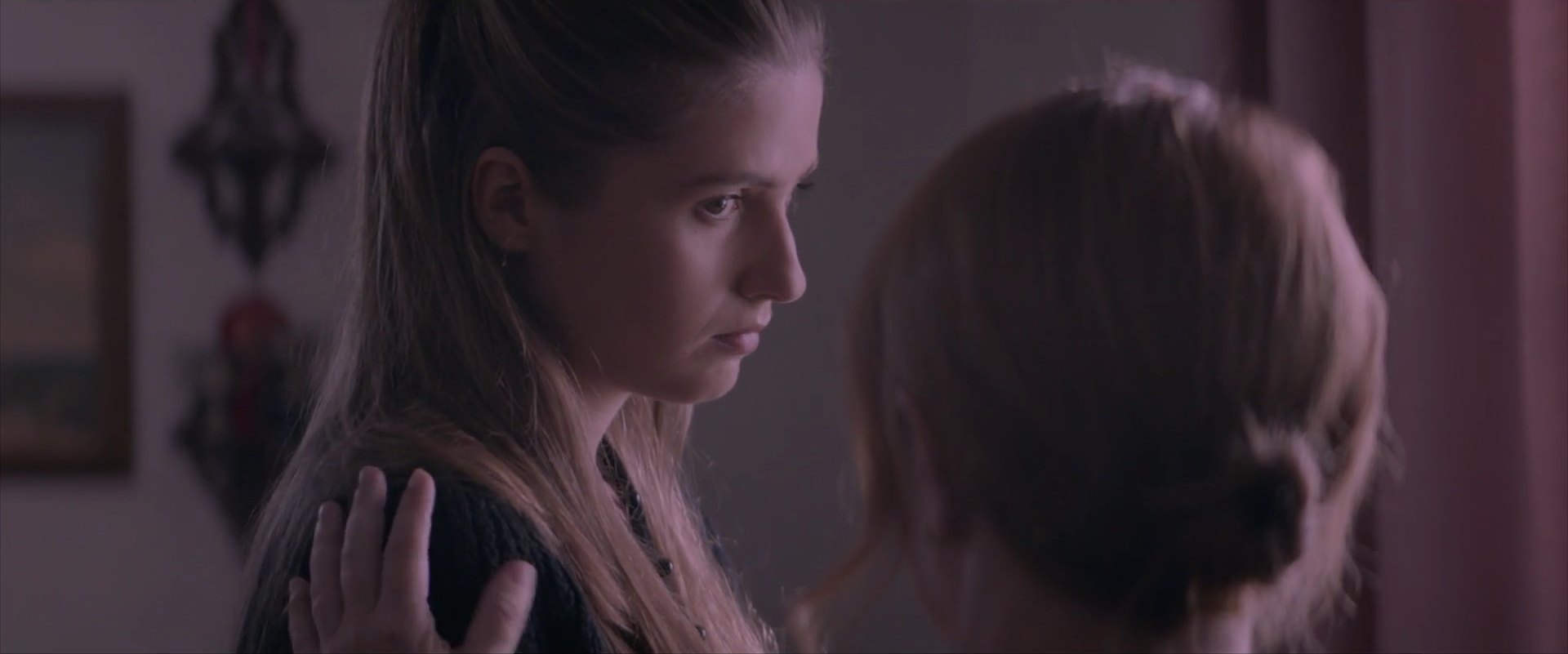 A young woman with long hair looks down while a hand rests on her shoulder, with another woman's head in the foreground.