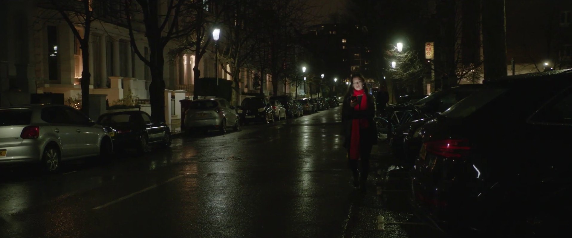 A woman in a red scarf walks down a dark, wet London street at night, looking at her phone.
