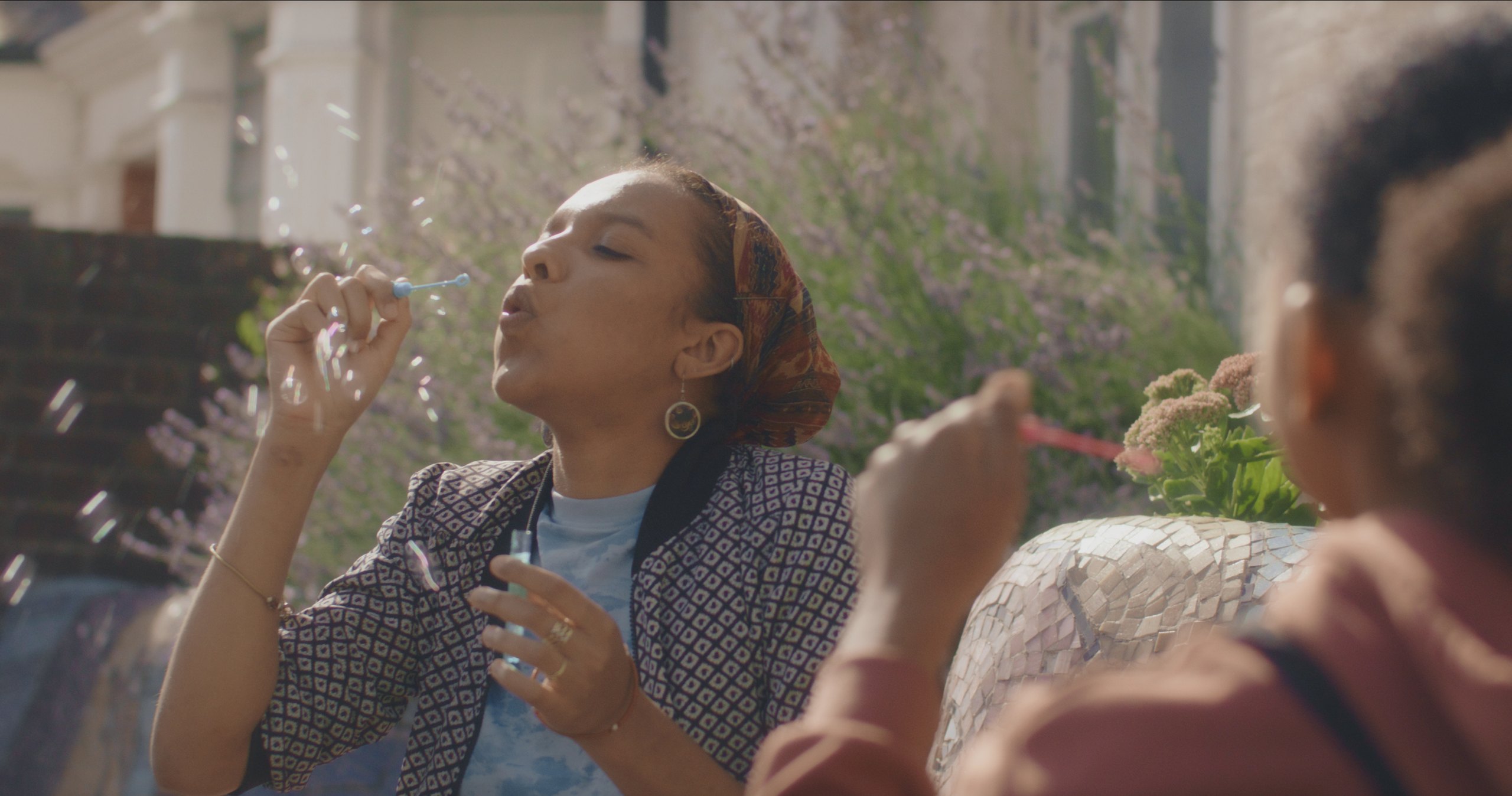 A woman in a patterned headscarf blows bubbles outdoors in sunlight, with a girl in the foreground doing the same.