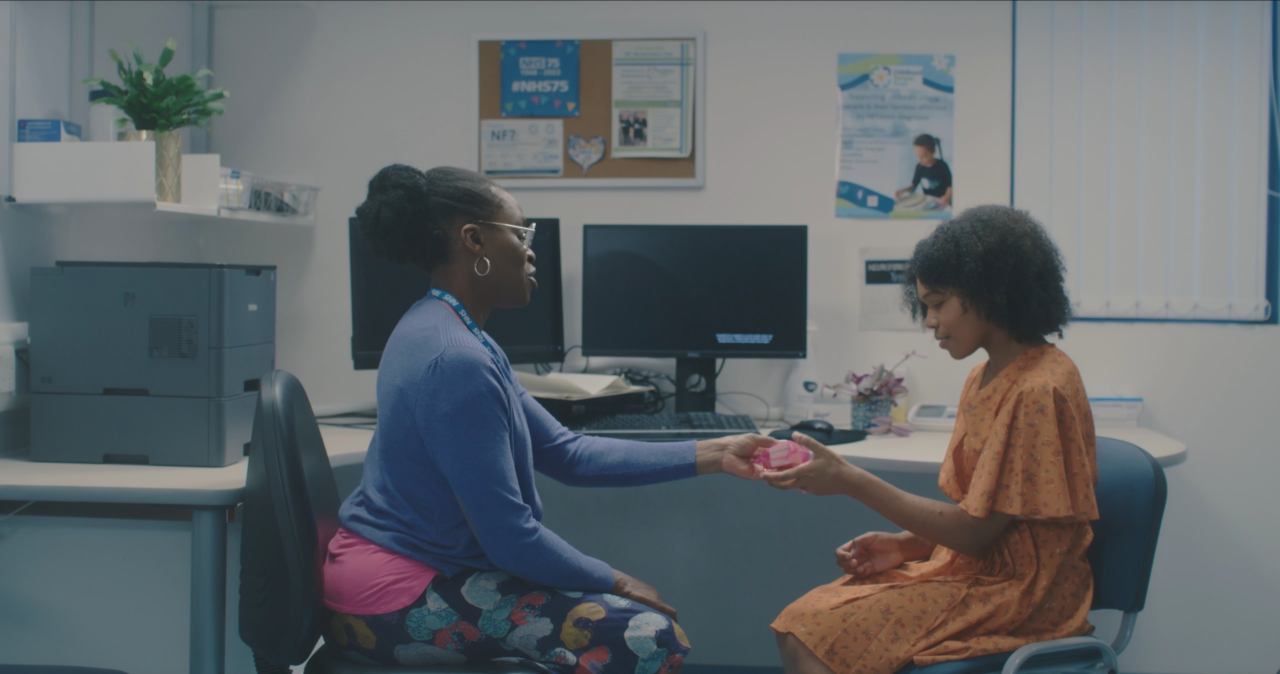 A nurse in a blue cardigan hands a small pink case to a teenage girl in an orange dress in a clinic office.
