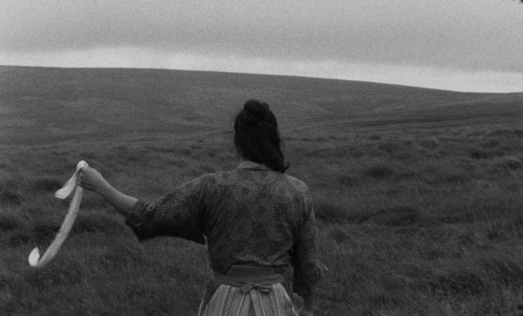 A woman seen from behind in a black-and-white moorland, holding a long white ribbon in one outstretched hand.
