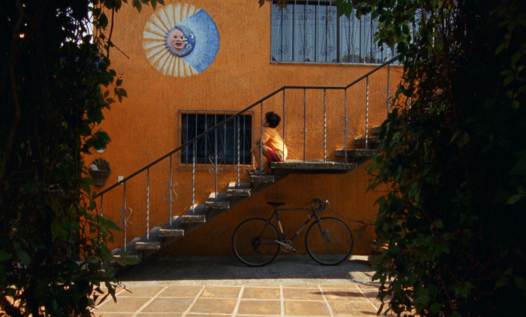 A boy crouches on a concrete staircase against an orange wall with a sun-and-moon mural, a bicycle leaning below.