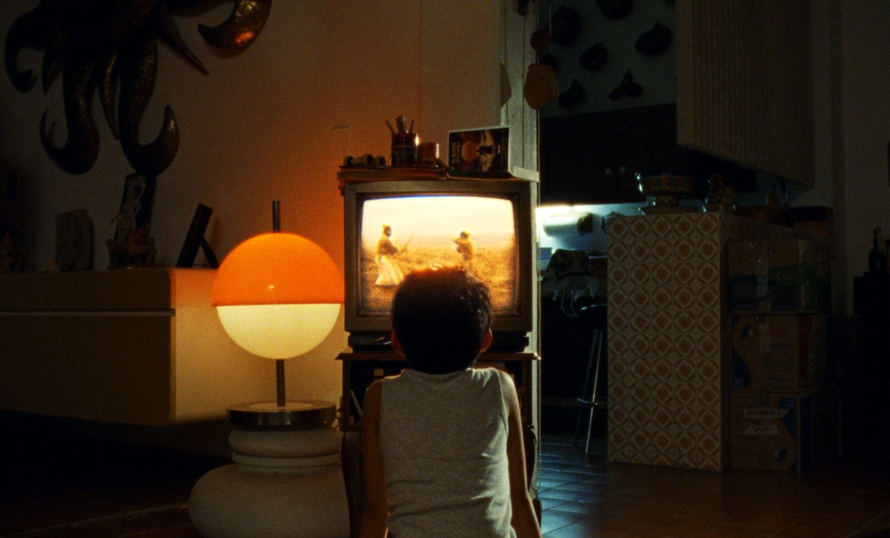 A child seen from behind watches an old television in a dim room, lit by an orange lamp.