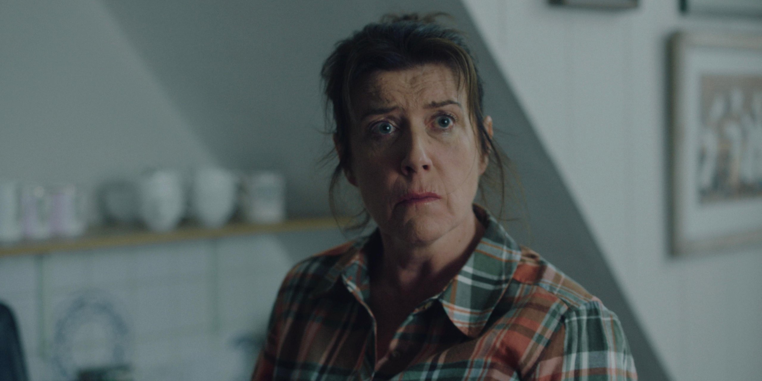 Close-up of a woman in a plaid shirt with wide, frightened eyes in a pale kitchen.