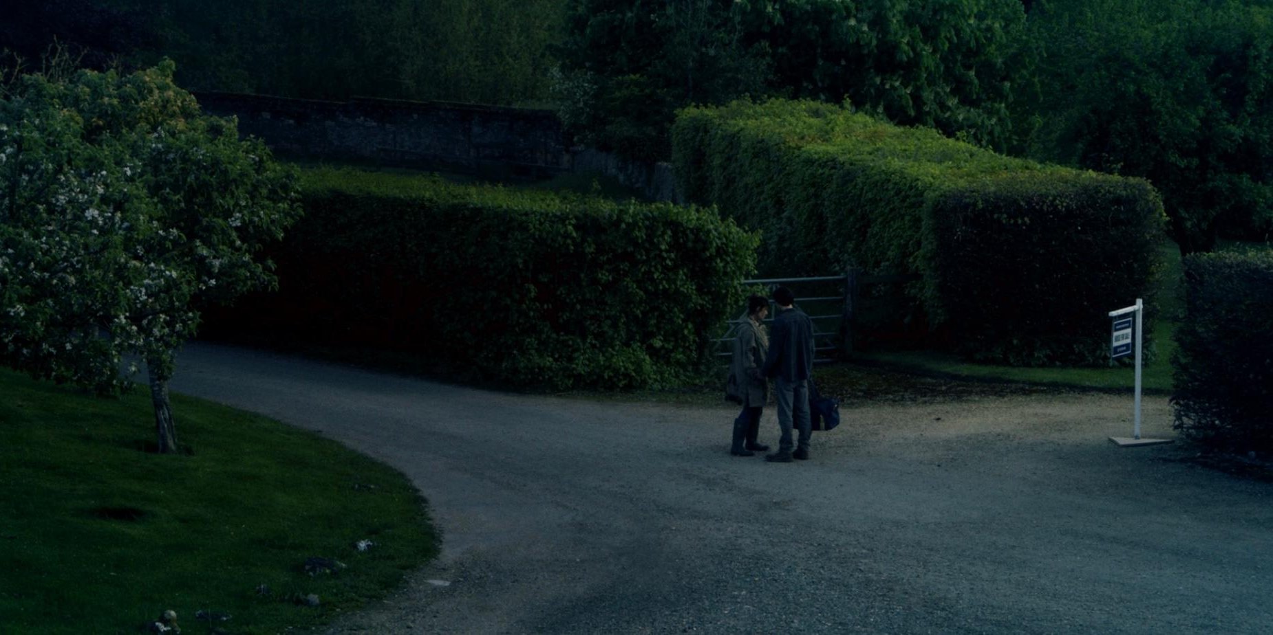 Two people stand close together at a gateway on a gravel drive between tall hedges at dusk, with a for-sale sign beside them.