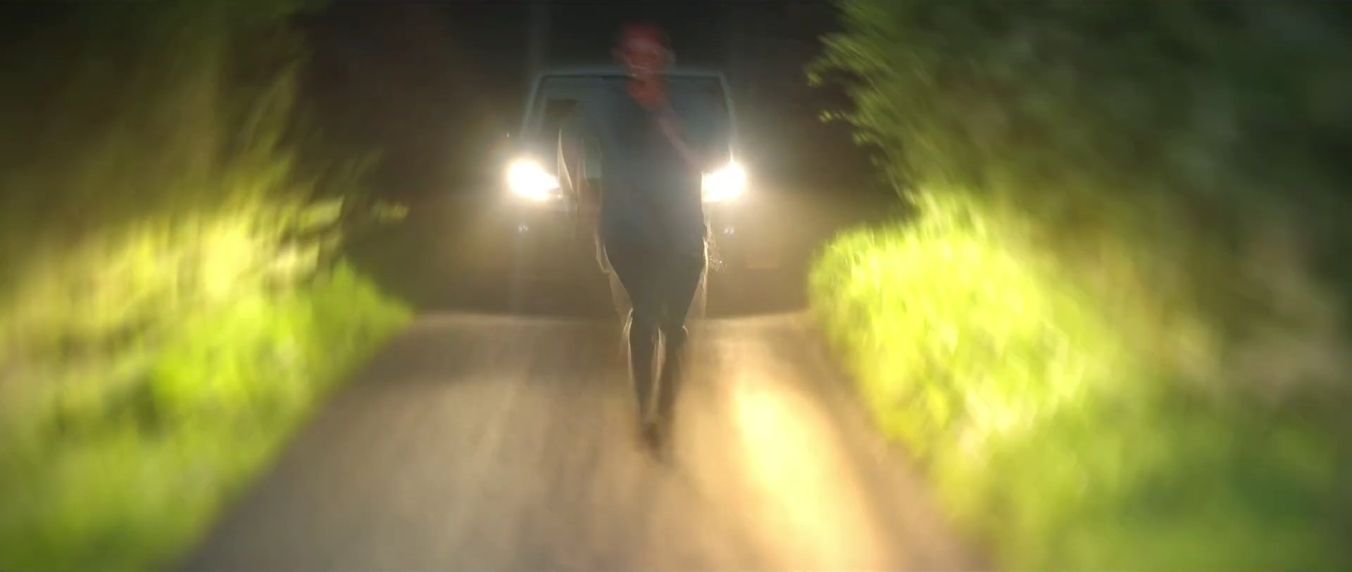 A blurred, dark figure runs along a lane toward a vehicle with its headlights on, hedges streaking past.
