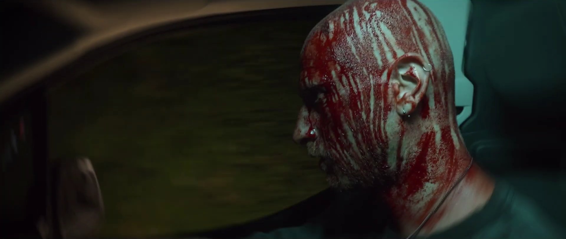 Profile of a bald man with blood across his head and face, with a motion-blurred blur passing in front.