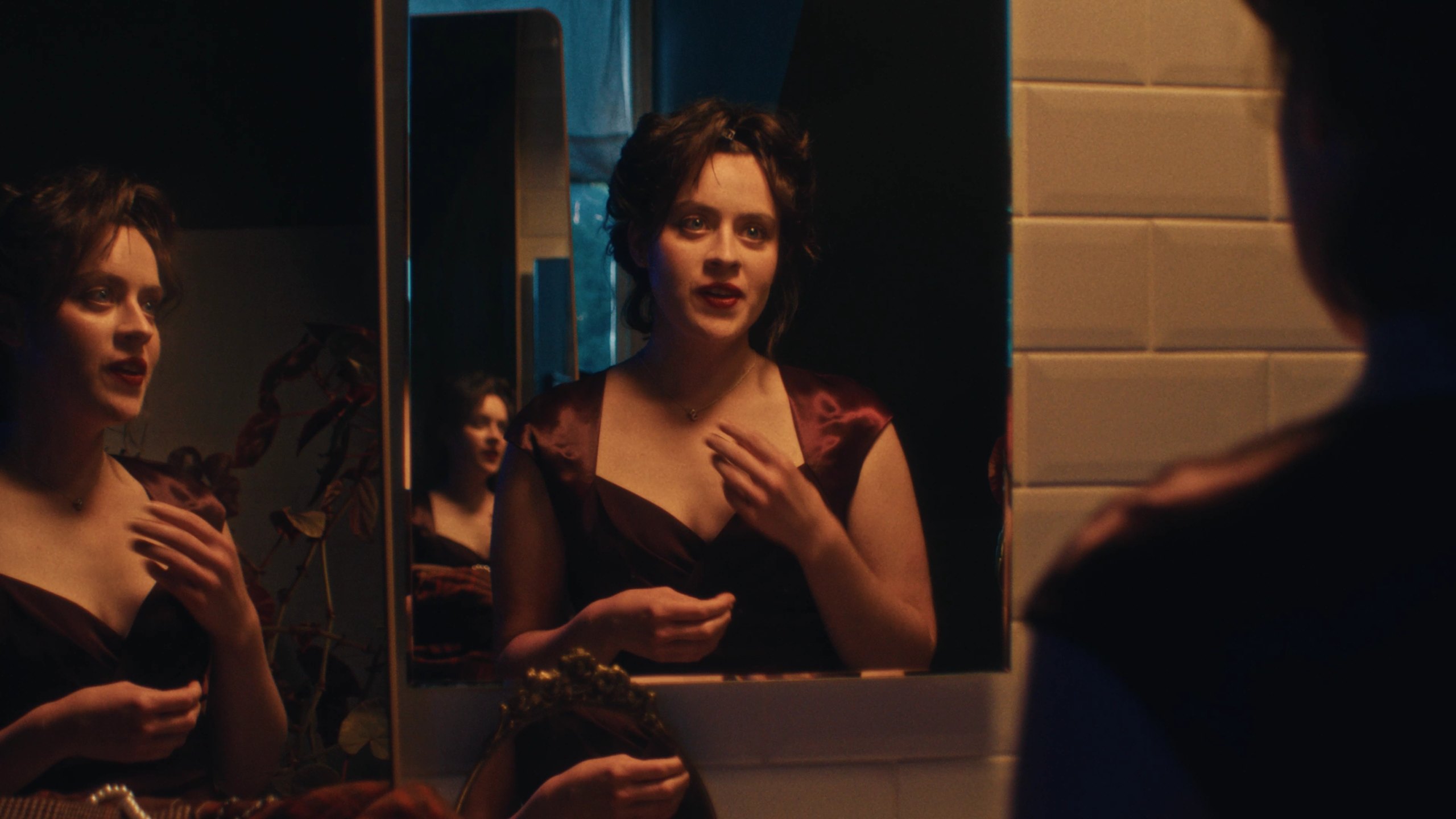 A woman in a dark red dress speaks to herself in a bathroom mirror, her reflection repeated in a second mirror.