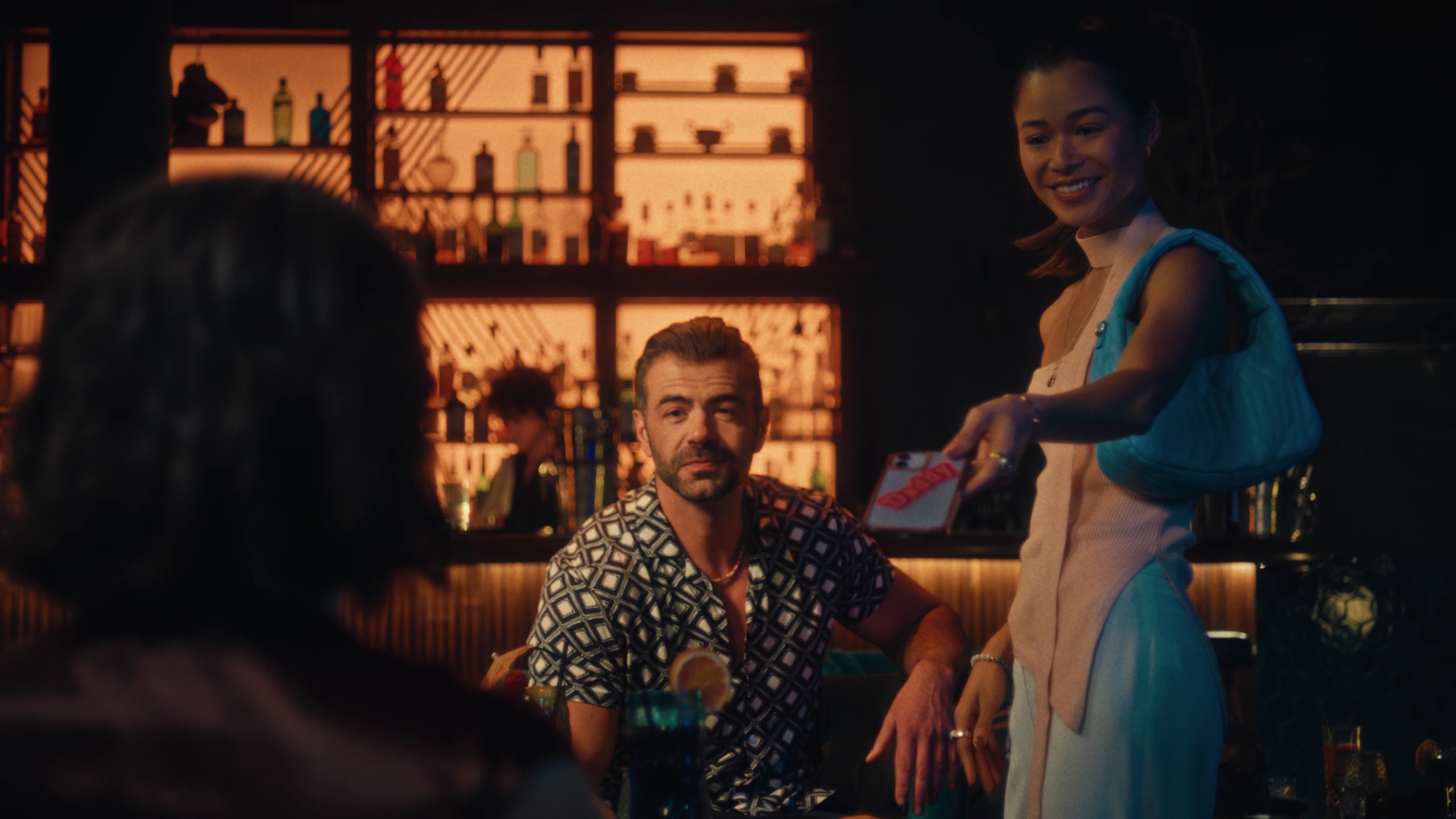 A man in a patterned shirt sits at a bar looking up at a smiling woman in a blue shoulder bag who holds out a phone.