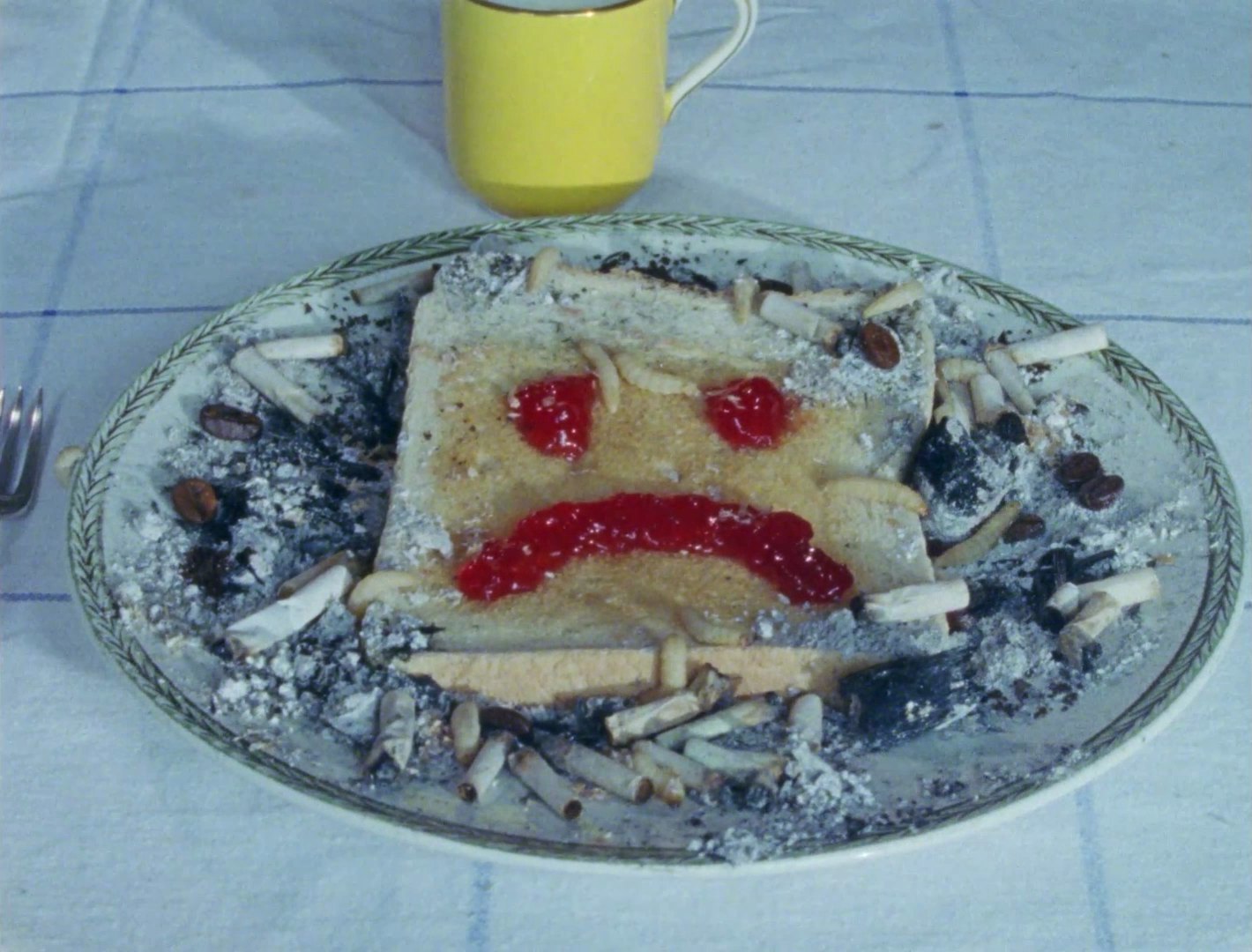 A slice of toast on a plate with a jam sad face, surrounded by cigarette butts, ash, coffee beans and maggots, beside a yellow mug.