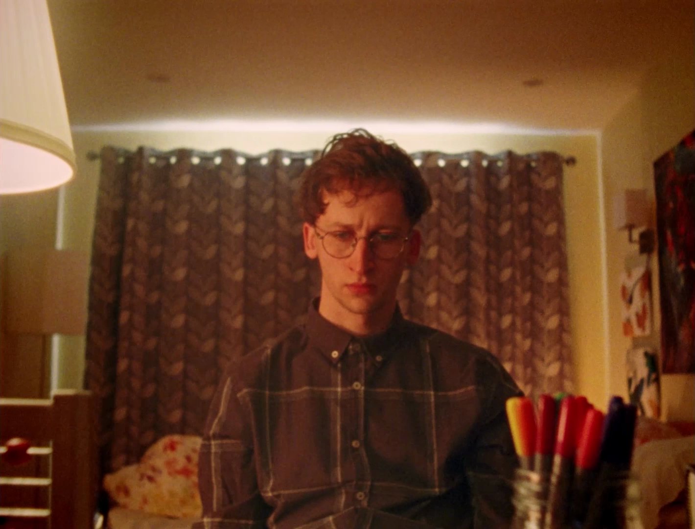 A young man with glasses and tousled hair sits in a warmly lit bedroom in front of patterned curtains, with felt-tip pens in the foreground.