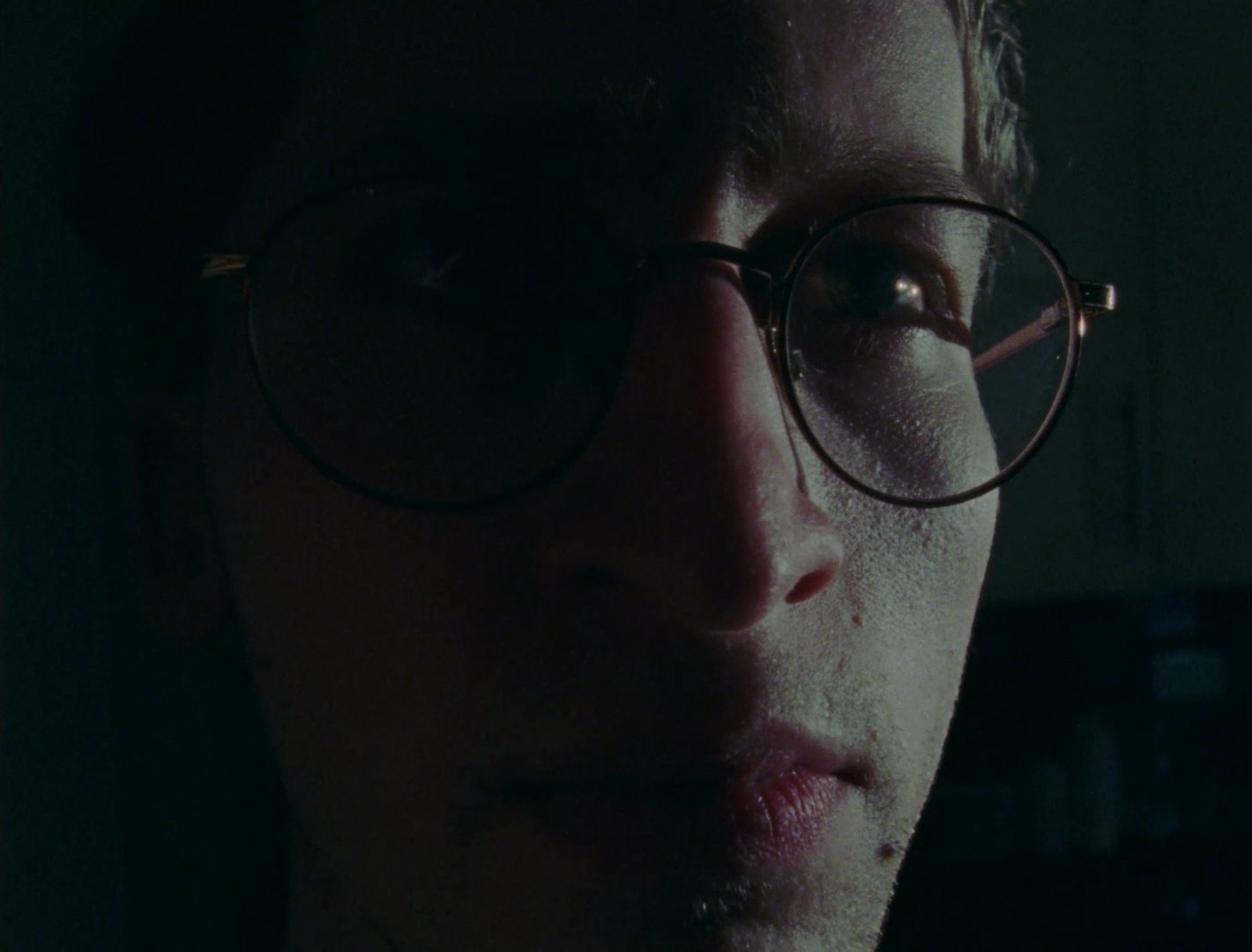 A close, half-lit portrait of a man in round glasses, his face partly in shadow.