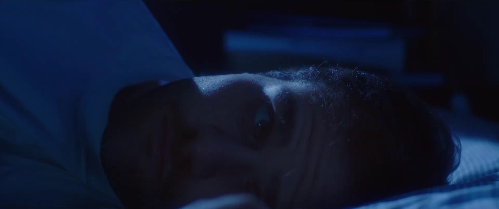 A man lies on his side in bed, lit in deep blue, his eye open in the dark.