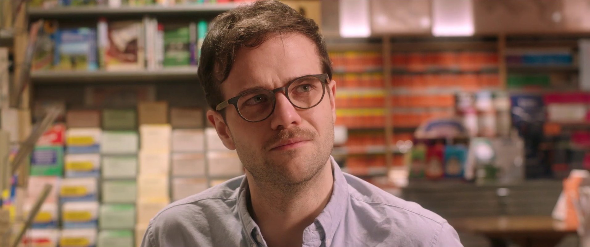 A man with glasses and a moustache looks off to the side in a shop with shelves of colourful packages behind him.